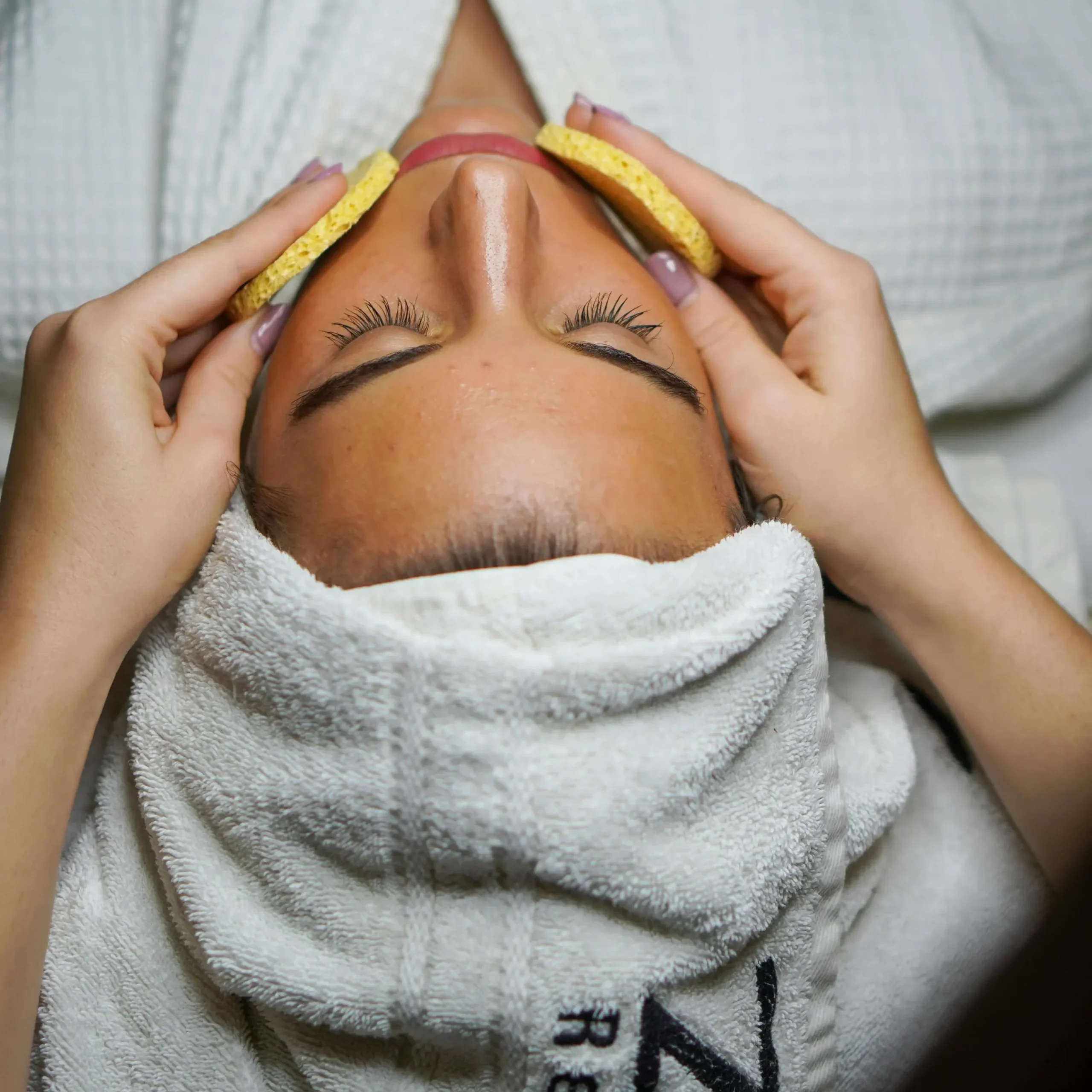Woman lying with eyes closed during a spa facial, wrapped in a white robe and towel, while a therapist gently massages her face with yellow sponges under soft lighting.