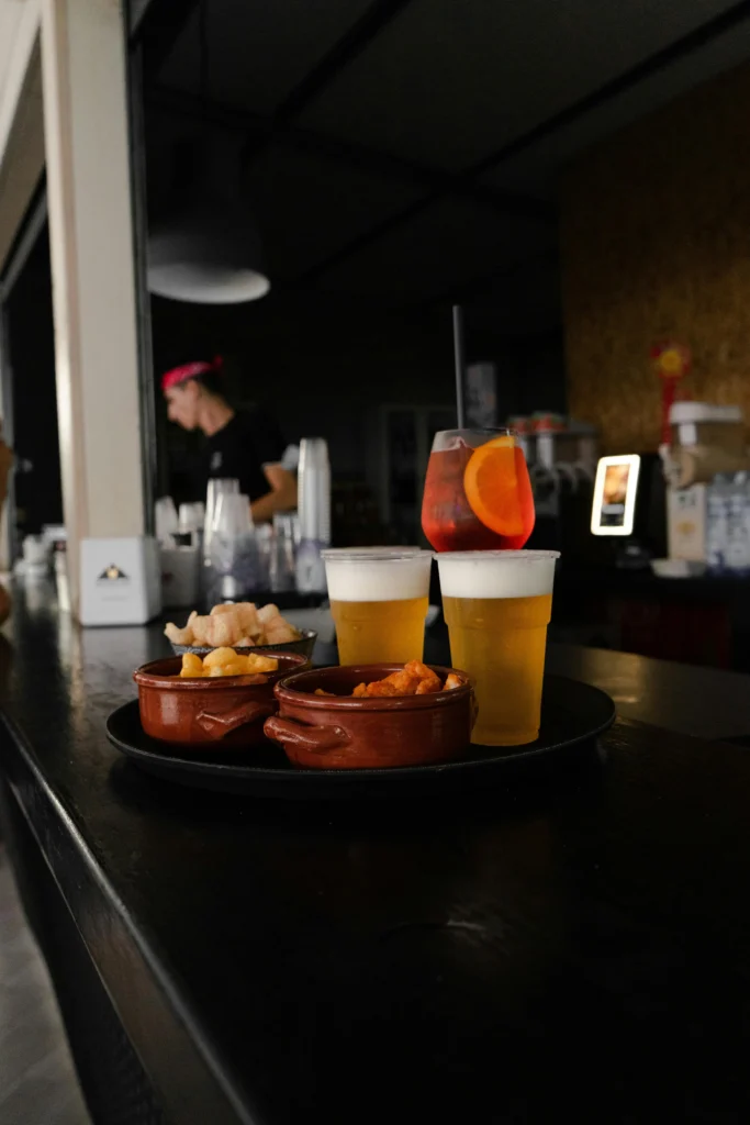 A tray with two glasses of beer and a colorful cocktail, accompanied by two small bowls of snacks, in a cozy bar setting.
