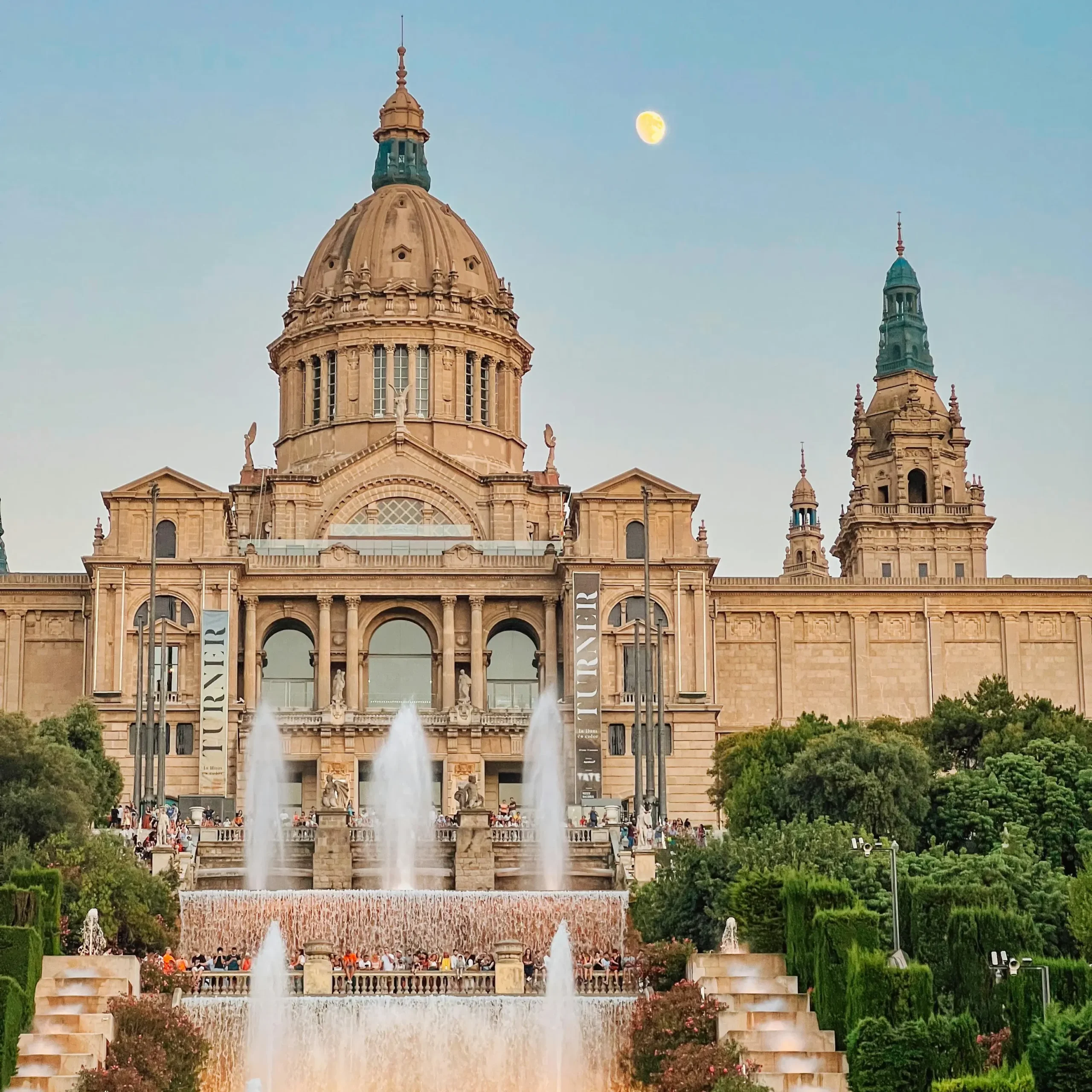View of the National Art Museum of Catalonia in Barcelona, with cascading fountains in the foreground, visitors enjoying the area, and the moon visible in the clear evening sky.