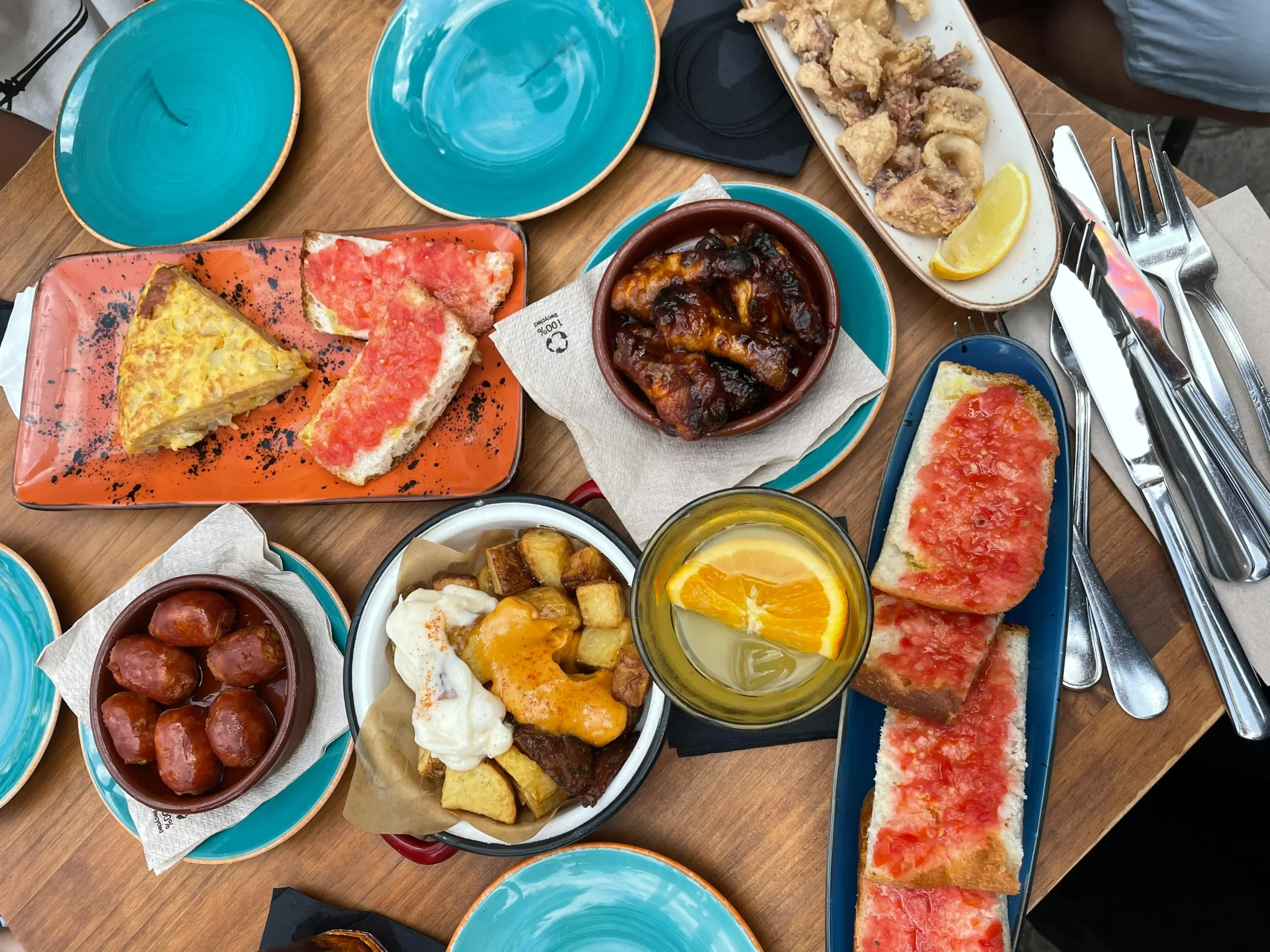 Selection of Spanish tapas including tortilla, chorizo, patatas bravas, pan con tomate, grilled meat, and fried calamari served on a wooden table.