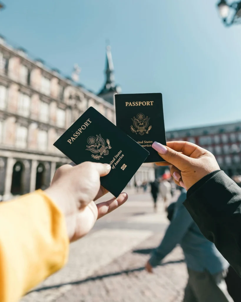 Two hands holding United States passports in a vibrant city square with historic buildings in the background.