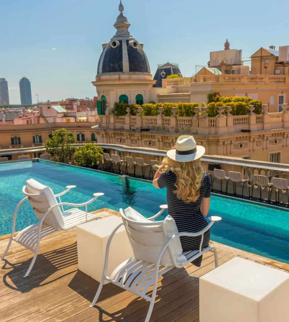 Femme portant un chapeau de soleil se détendant au bord d’une piscine de luxe sur un toit, avec des vues panoramiques sur l’architecture historique de Barcelone par une journée ensoleillée.