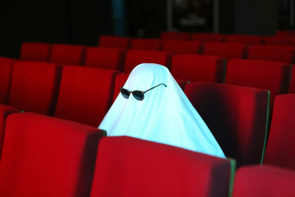 Persona disfrazada de fantasma con sábana blanca y gafas de sol sentada en una butaca roja de un teatro en Barcelona.