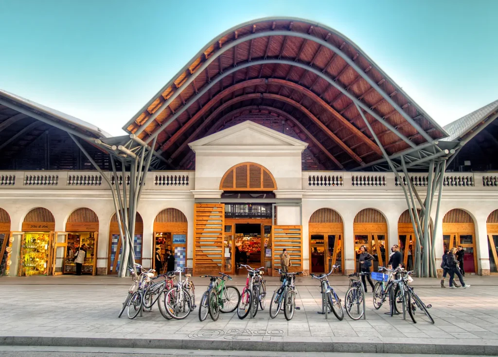 Vista exterior del Mercat de Santa Caterina en Barcelona, con bicicletas estacionadas frente al edificio y personas caminando por la plaza. La arquitectura del mercado destaca por su techo ondulado y estructura metálica.