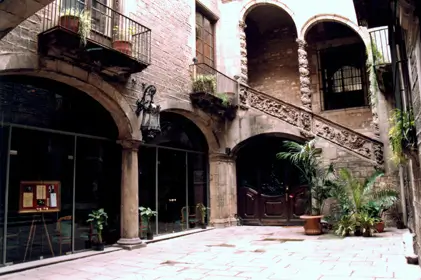 Una vista del Palau Dalmases, un edificio histórico en el Barrio Gótico de Barcelona, con su característico patio interior y arquitectura medieval. El lugar está adornado con plantas y columnas de piedra, creando una atmósfera única y acogedora.