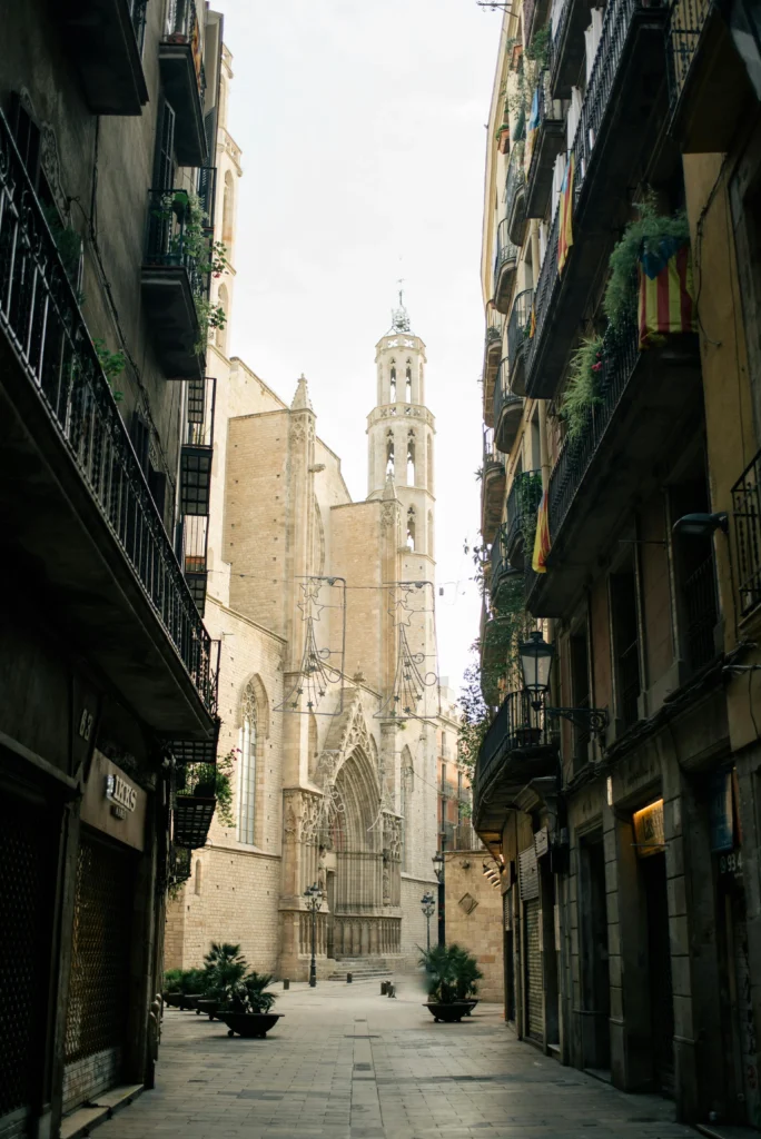 Vista de la Iglesia de Santa María del Mar, ubicada en el Barrio del Born, Barcelona. La imagen muestra una calle tranquila con balcones adornados y la impresionante torre de la iglesia al fondo.