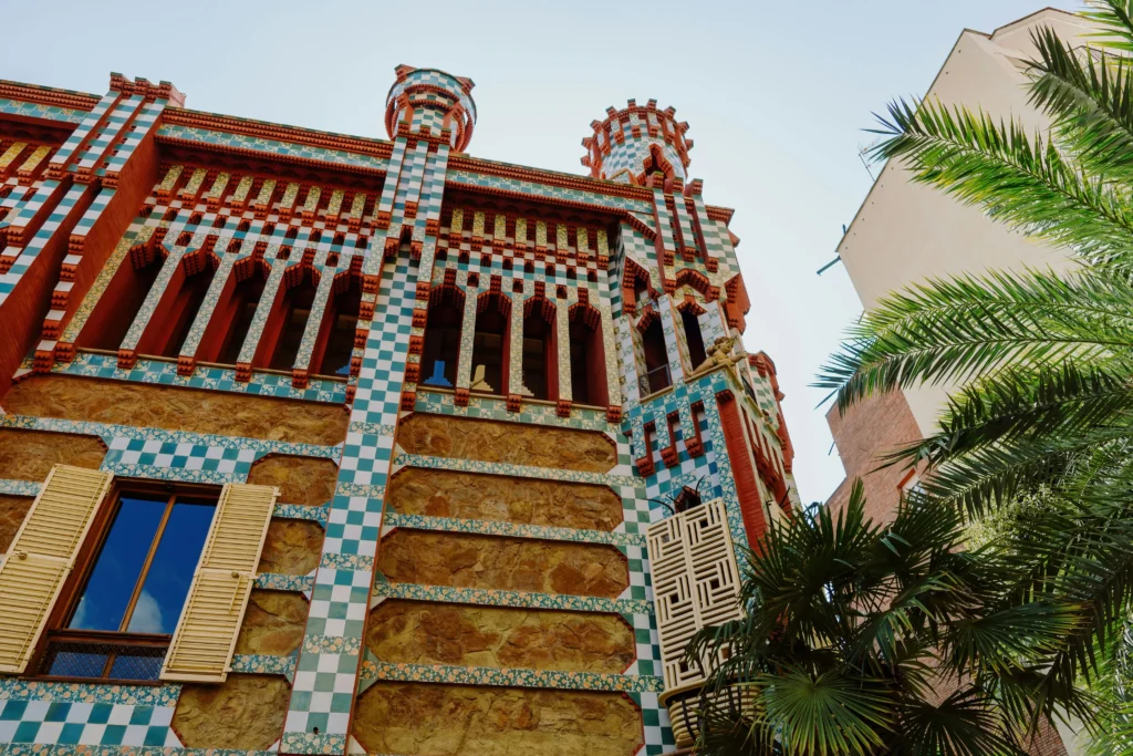 Fachada colorida de la Casa Vicens en Barcelona, diseñada por Antoni Gaudí, con cerámica verde, roja y blanca, rodeada de palmeras y edificios modernos.