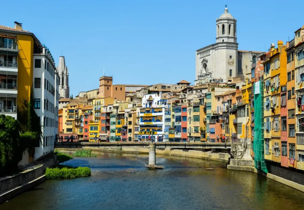 Fotografía diurna de las casas coloridas a orillas del río Onyar, con la Catedral de Girona de fondo y un puente peatonal cruzando el río.