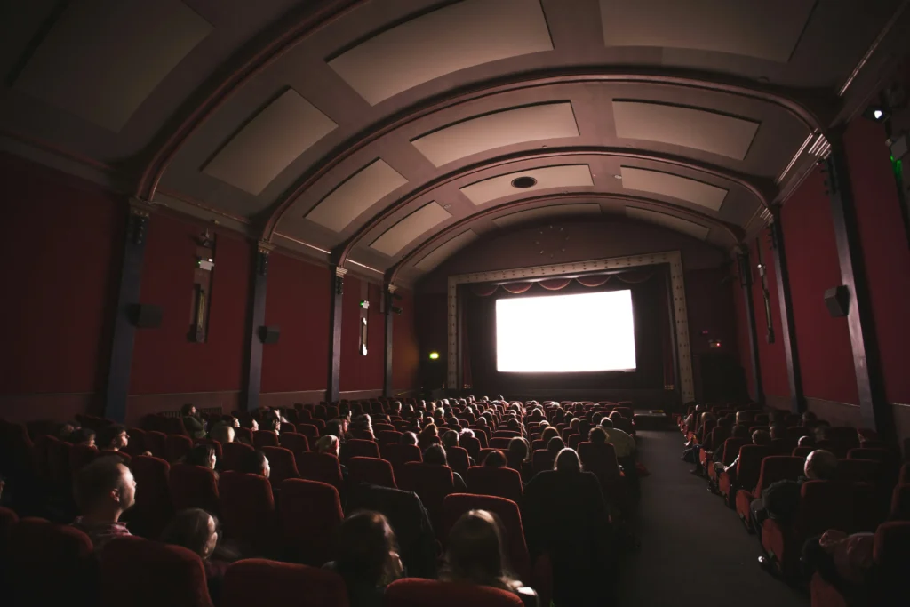 Interior de una sala de cine clásica con techo abovedado, asientos rojos y espectadores mirando una película en pantalla grande iluminada. Ambiente acogedor y nostálgico.