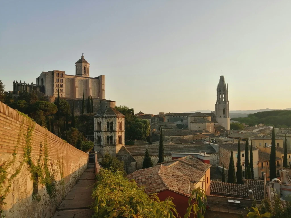 Vista al atardecer de Girona desde las antiguas murallas, con la Catedral de Santa María al fondo, el campanario de Sant Feliu y tejados históricos entre cipreses.