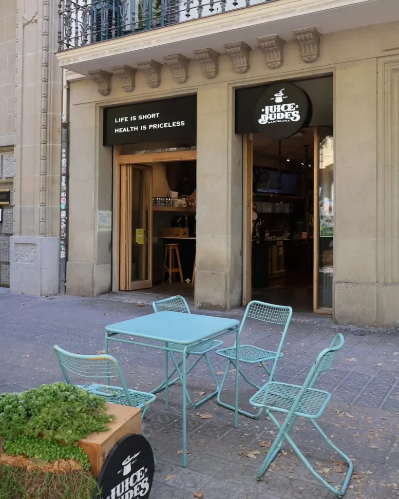 Fachada de Juice Dudes con mesas turquesa en la terraza, ubicada en una calle peatonal de Barcelona con ambiente relajado y saludable.