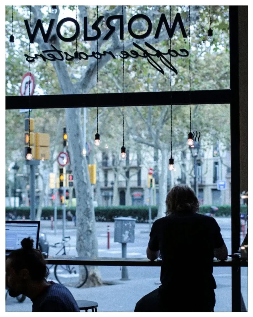 Interior de Morrow Coffee con vista a la calle en Barcelona, iluminación tenue y personas disfrutando de su café en la barra frente al ventanal.