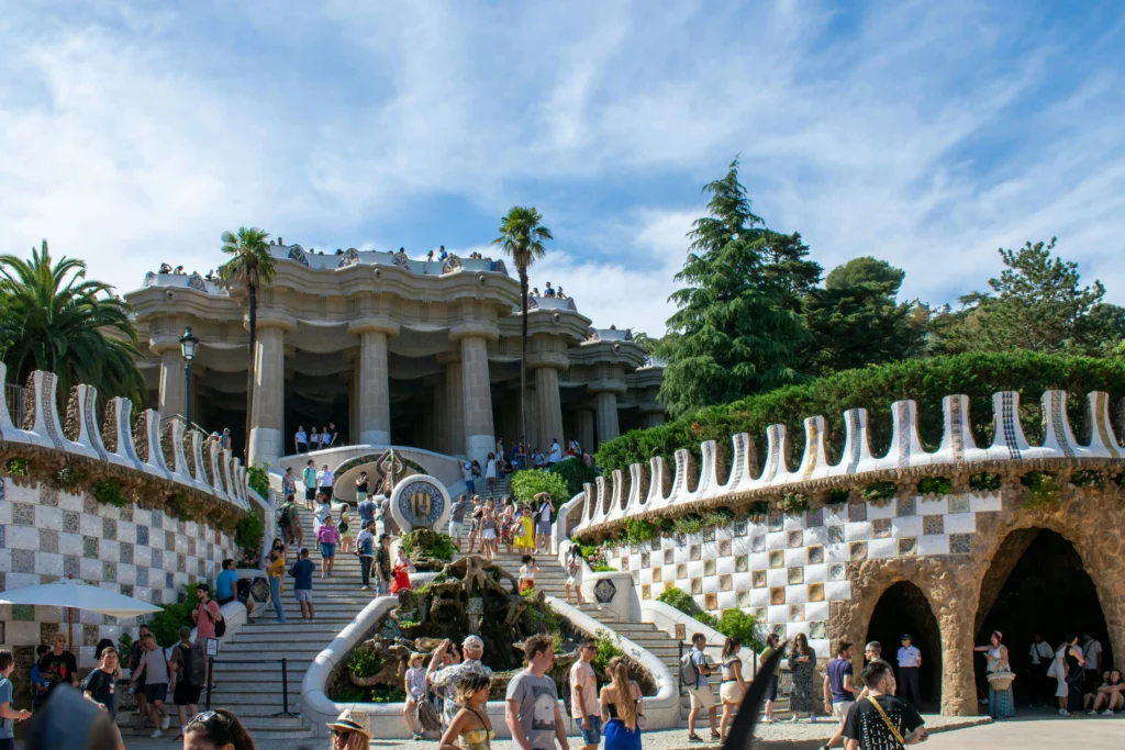 The main staircase of Park Güell in Barcelona filled with tourists, showing the iconic mosaic dragon, curving walls, and Doric columns of the Hypostyle Room surrounded by lush greenery.