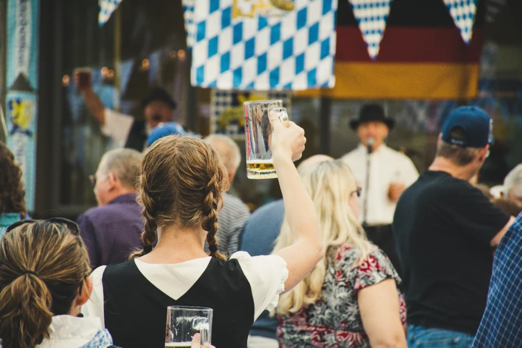 A group of people celebrating at an Oktoberfest event, with one person raising a beer mug in a toast, while others enjoy the lively atmosphere. The background features German flags and festive decorations.