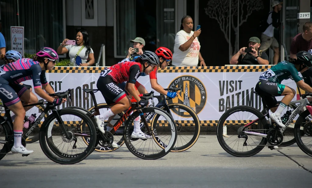 Una carrera de ciclismo en Baltimore, con varios ciclistas en acción, incluyendo uno con un jersey rojo y otro con uno verde. Los espectadores observan y toman fotos a lo largo del recorrido. Al fondo se ve un banner de "Visit Baltimore