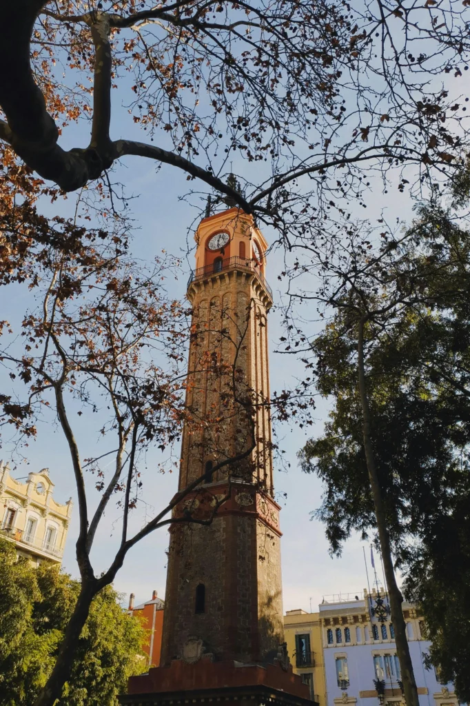 Torre del Reloj de Sant Andreu de Palomar en Barcelona, rodeada de árboles desnudos en otoño, con cielo despejado y edificios modernistas al fondo.