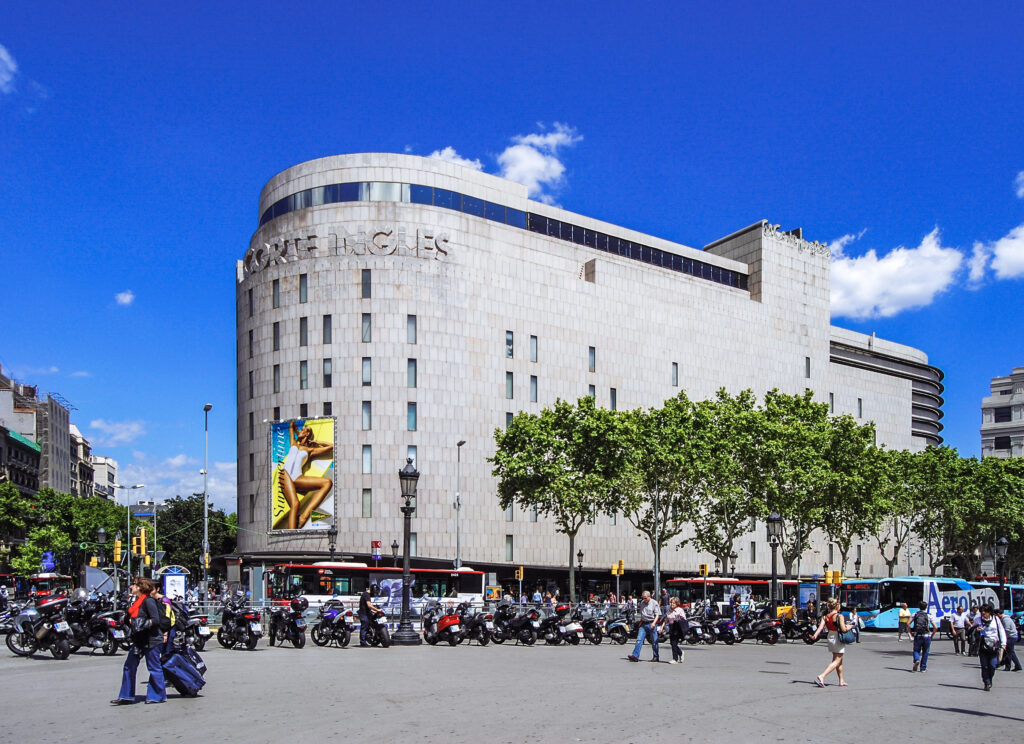 Fachada principal de El Corte Inglés en Plaça Catalunya, Barcelona, con peatones y vehículos en una zona comercial muy transitada.