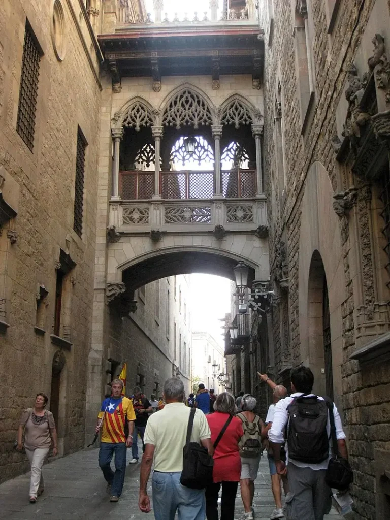 Imagen del Carrer del Bisbe con su famoso puente neogótico sobre la calle, rodeado de edificios históricos y con varios visitantes caminando por el pasaje.