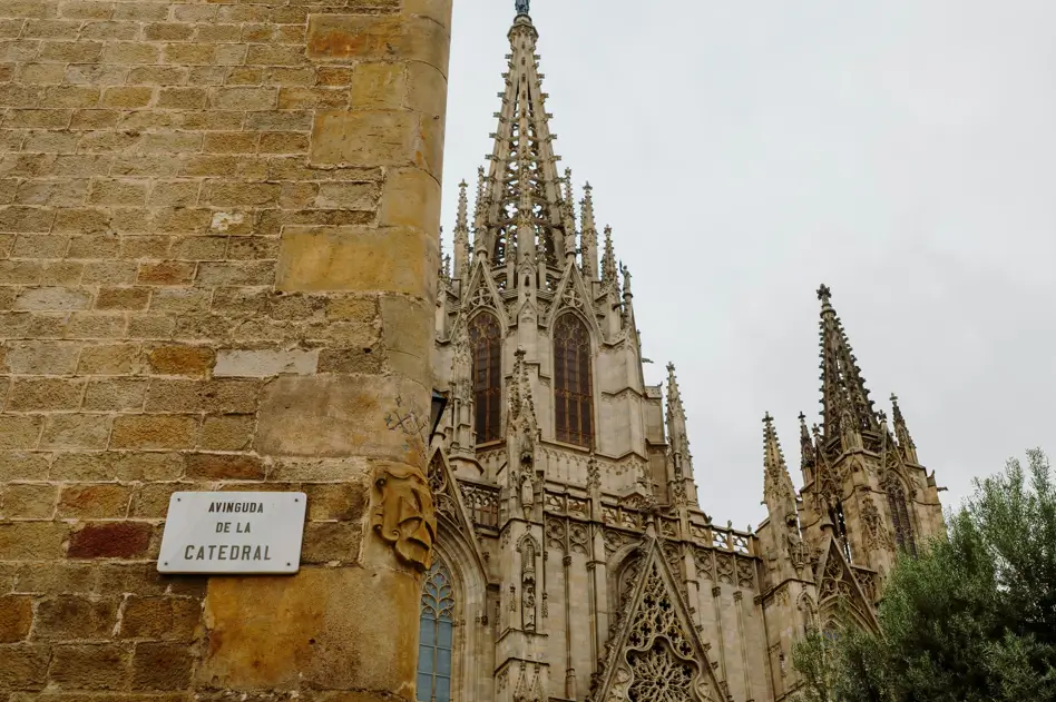 Vista parcial de la Catedral de Barcelona con sus torres góticas, tomada desde la Avinguda de la Catedral, junto a una pared de piedra con el rótulo de la calle.