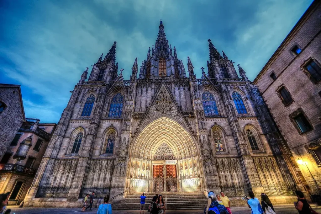 Fachada gótica de la Catedral de Barcelona iluminada al anochecer, vista desde la plaza del Barrio Gótico con varios visitantes observando y haciendo fotos.