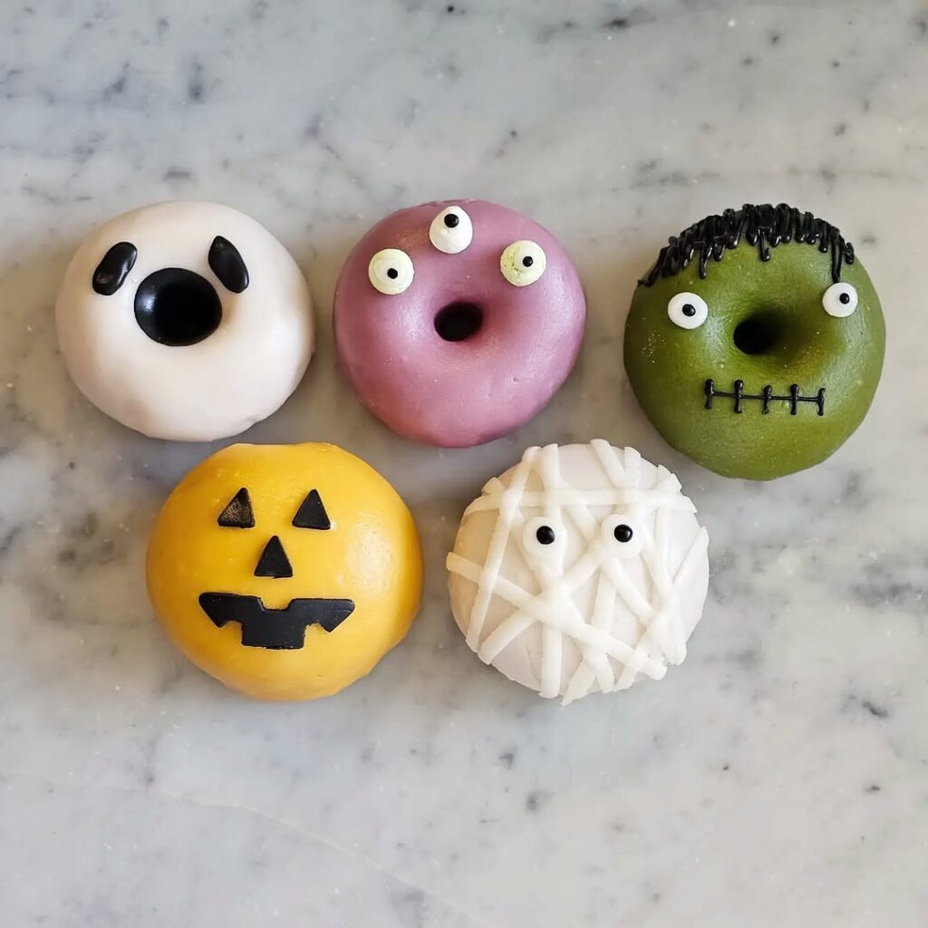 Пять пончиков, украшенных в стиле Хэллоуина, на мраморной поверхности: один в виде призрака, один розовый с тремя глазами, один зелёный в стиле Франкенштейна, один жёлтый — тыква и один белый в виде мумии.