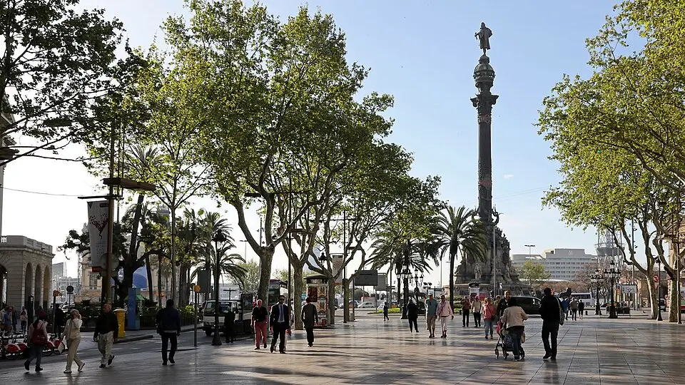 Paseo de La Rambla en Barcelona con gente caminando bajo los árboles y el Mirador de Colom al fondo, cerca del puerto.