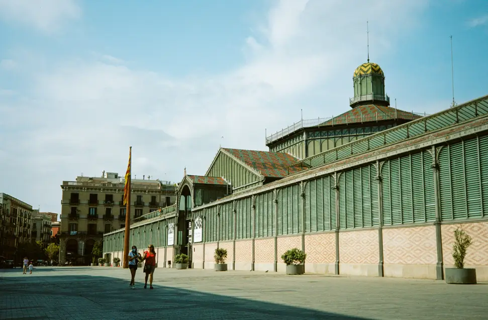 Vista amplia del exterior del Mercat del Born en Barcelona, con su estructura modernista de hierro y la plaza abierta donde pasean varios visitantes.