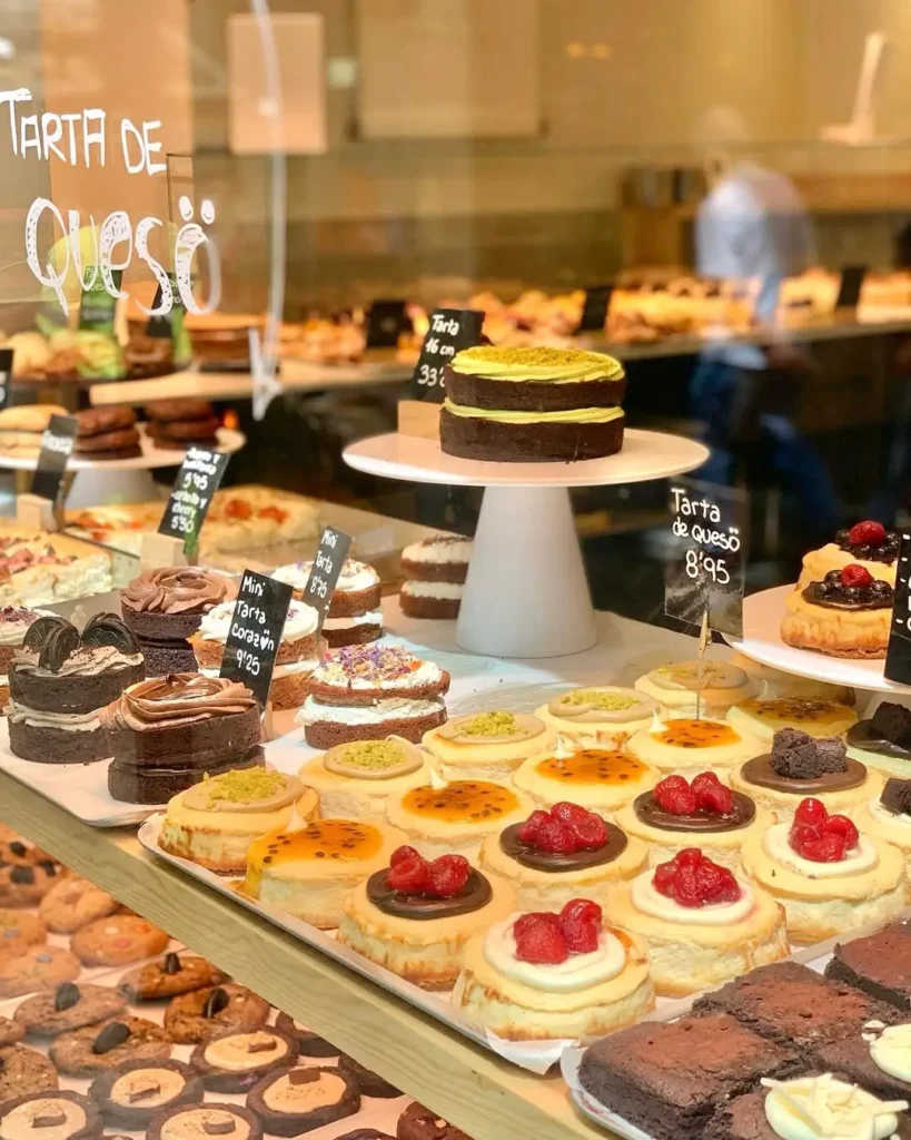 Витрина кондитерской в Барселоне, полная чизкейков, мини-пирожных и домашнего брауни, украшенных фруктами и шоколадом, с ценниками.