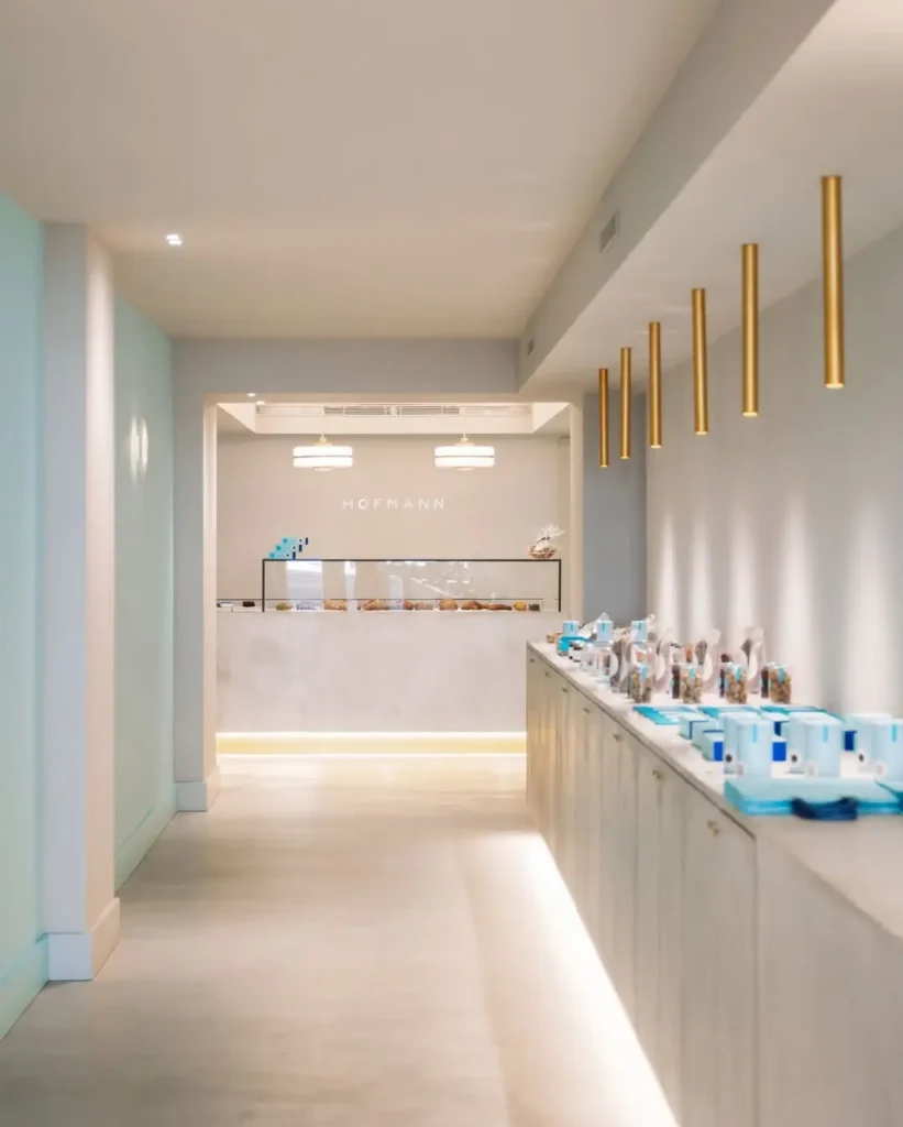 Элегантный и светлый коридор кондитерской Hofmann в Барселоне с минималистичным дизайном, мягкими тонами и витриной на заднем плане с гастрономическими продуктами и деталями в синем и золотом.