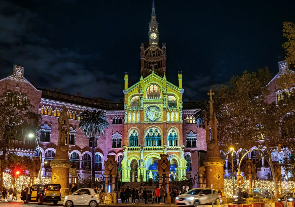 Fachada principal del Recinte Modernista de Sant Pau iluminada por luces de colores durante la noche, con detalles arquitectónicos modernistas, palmeras y visitantes frente a la entrada.