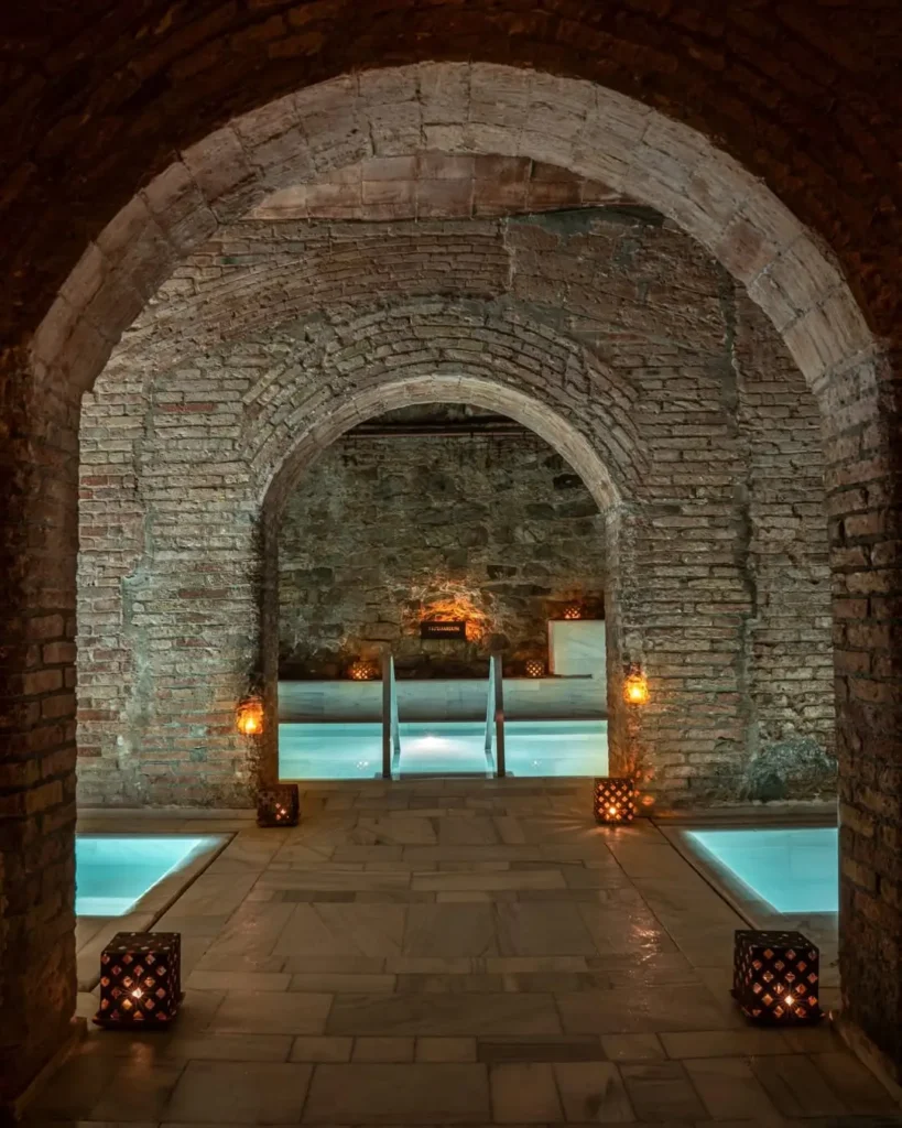 Вид на термальные бассейны хаммама в Барселоне, окруженные кирпичными стенами и масляными лампами, создающими атмосферу расслабления и очищения.