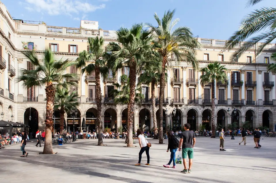 Vista de la Plaça Reial en el Barrio Gótico, con palmeras altas, terrazas llenas de gente y visitantes paseando por la plaza.
