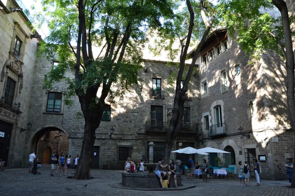 Imagen de la Plaça Sant Felip Neri en Barcelona, con su fuente central, árboles que proyectan sombras sobre las fachadas históricas y personas descansando en un ambiente tranquilo.