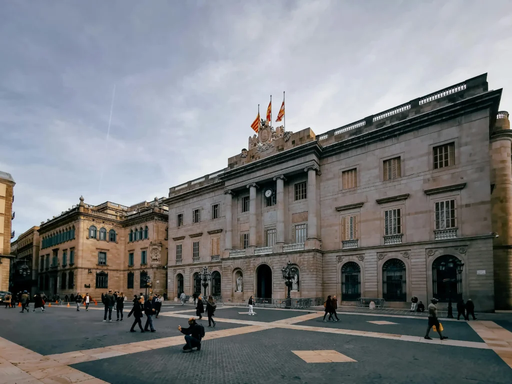 Vista amplia de la Plaça Sant Jaume en el Barrio Gótico, con el Ayuntamiento de Barcelona al fondo y varias personas caminando por la plaza.