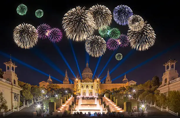 Vista nocturna del Palacio Nacional de Montjuïc iluminado, con fuegos artificiales de varios colores en el cielo y luces proyectadas sobre la explanada, creando un ambiente festivo y multitudinario.