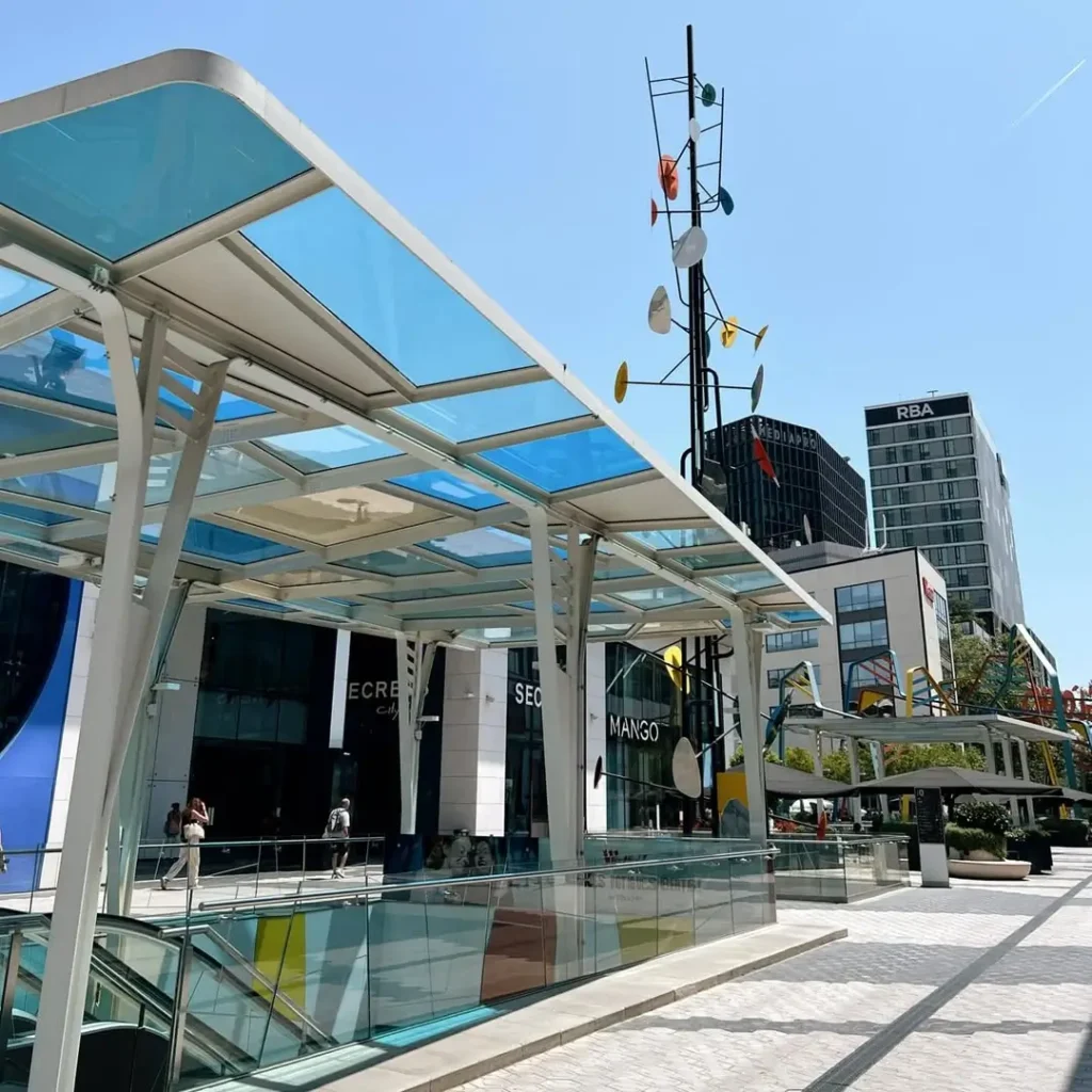 Vista exterior del Centro Comercial Westfield Glòries en Barcelona, con estructuras de cristal azul, escaleras de acceso y edificios modernos alrededor en un día soleado.