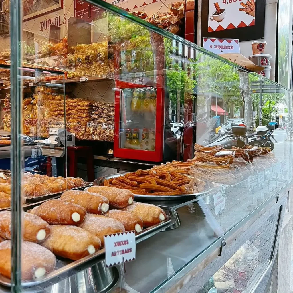 Витрина традиционной пекарни в Барселоне с подносами, наполненными кремовыми чуксами и свежими чуррос, рядом с полками, полными типичных каталонских сладостей.