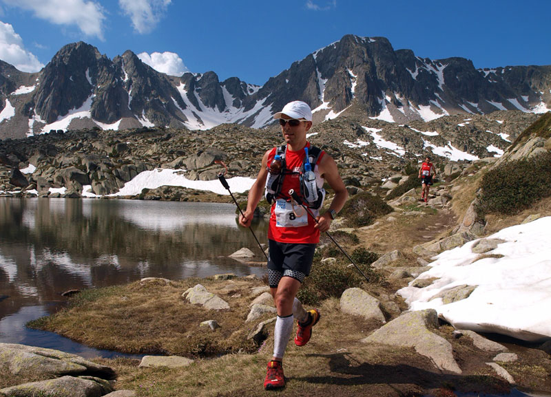Corredor de trail running avanzando por un sendero de alta montaña junto a un lago con nieve y picos rocosos al fondo, en un paisaje natural de Andorra.
