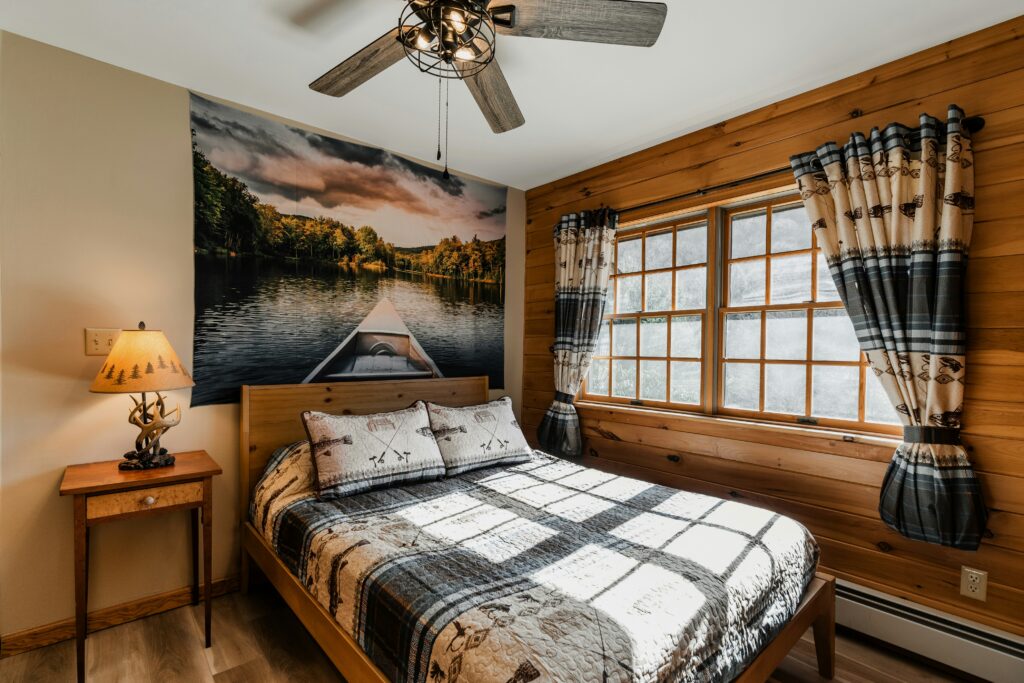 Habitación acogedora de estilo rústico con paredes de madera, cama decorada con textiles temáticos y un gran mural de un lago en la cabecera, iluminada por luz natural a través de ventanas de marco de madera.