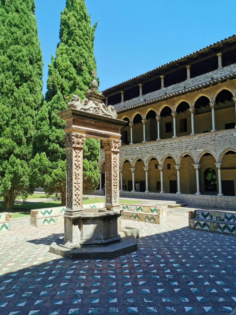 Pozo gótico en el claustro del Monasterio de Pedralbes de Barcelona, rodeado de arcos medievales, azulejos decorativos y jardines con cipreses bajo un cielo despejado.