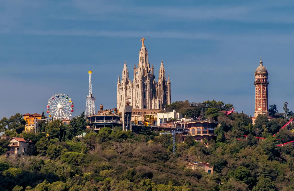 El Tibidabo de Barcelona con el Templo del Sagrat Cor al centro, la noria del parque de atracciones y la torre panorámica, rodeados de vegetación en la montaña.