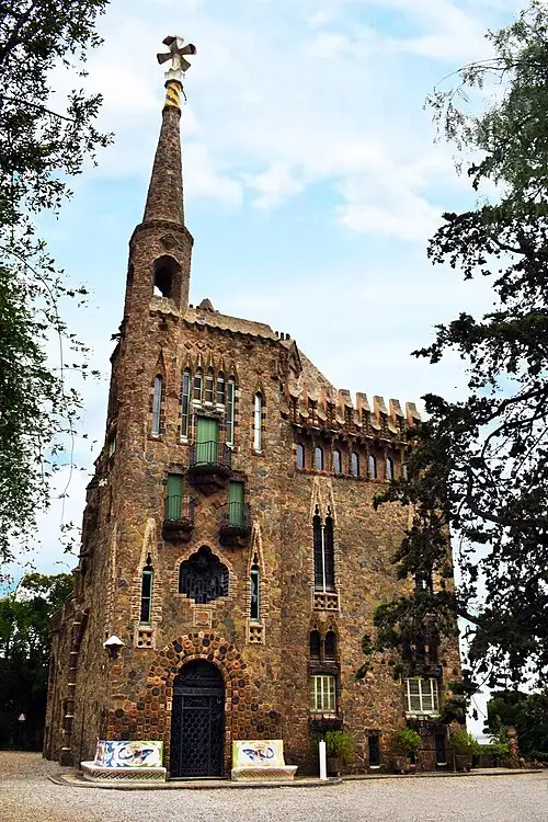 View of Gaudí’s Bellesguard Tower in Barcelona, featuring its stone façade, Gothic-style windows, and the distinctive tower topped with a cross, surrounded by greenery.