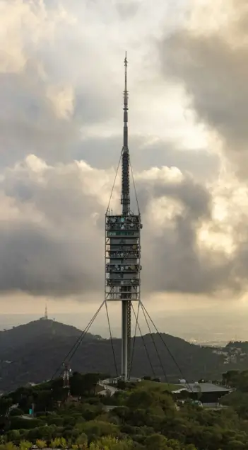 La Torre de Collserola en Barcelona vista desde la montaña, rodeada de nubes densas y luz de atardecer, con colinas y vegetación en primer plano.