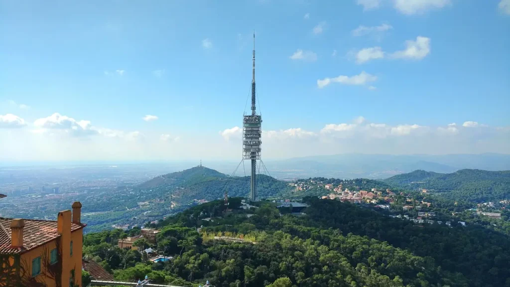 Vista panorámica de Barcelona con la Torre de Collserola en primer plano, rodeada de colinas verdes y cielo despejado.