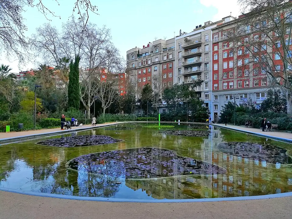 Pond at Turó Park in Barcelona, surrounded by gardens, trees, and residential buildings, with visitors strolling and enjoying the peaceful setting.