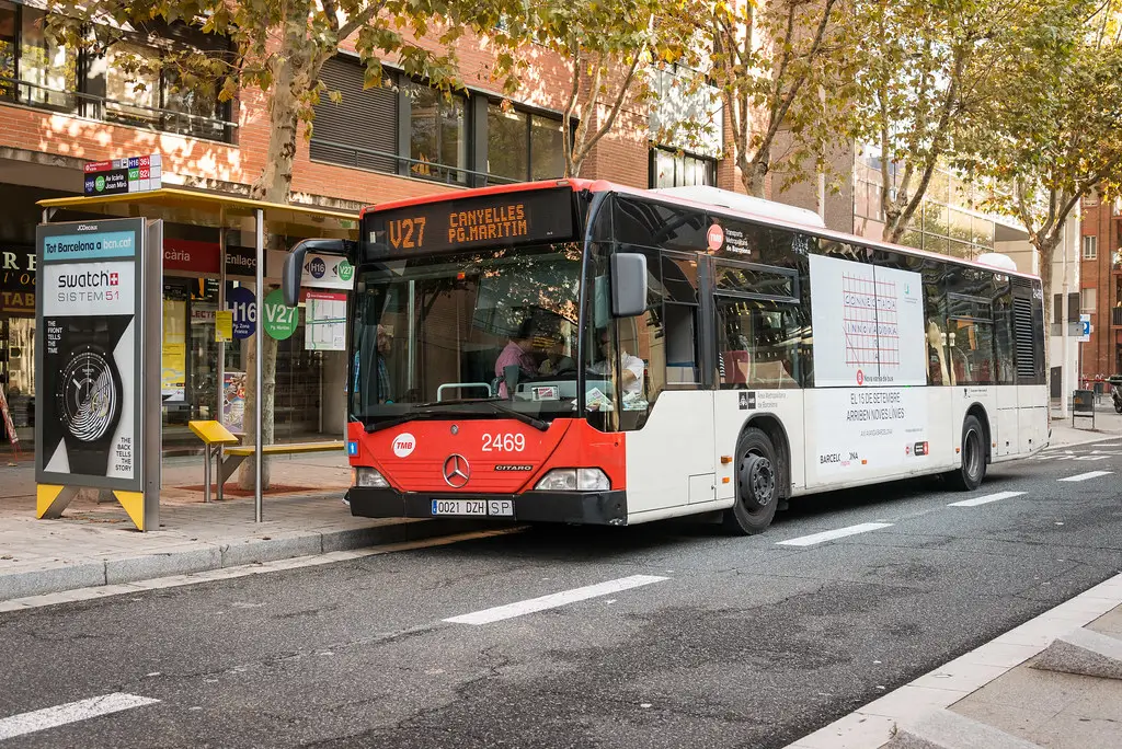 TMB city bus stopped at a street bus stop in Barcelona, with line signage and residential buildings nearby.
