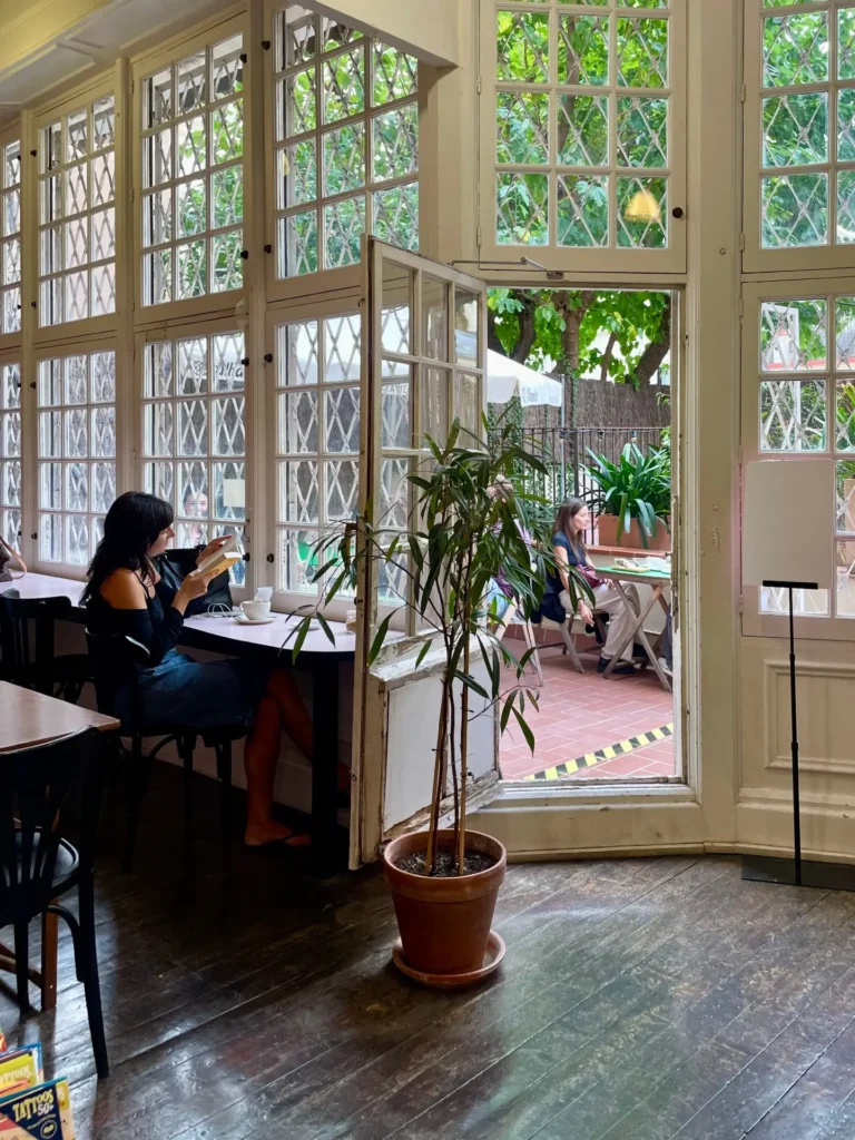 Interior de una cafetería luminosa en Barcelona, con grandes ventanales abiertos a un patio interior verde, mesas de madera y personas disfrutando tranquilamente de café y lectura.