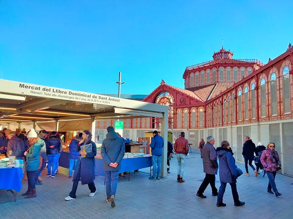Stalls at the Sant Antoni Sunday market selling books and collectibles, with visitors browsing outdoor stands and the modernist market building in the background, Barcelona