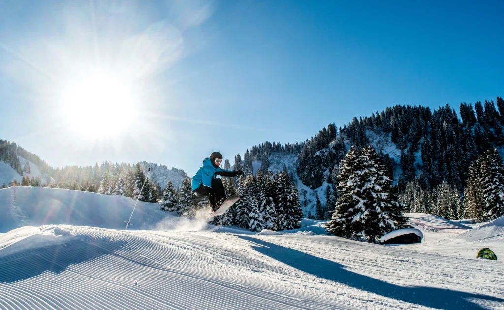 Snowboarder realizando un salto en una pista nevada rodeada de árboles, en un día soleado de invierno en la montaña