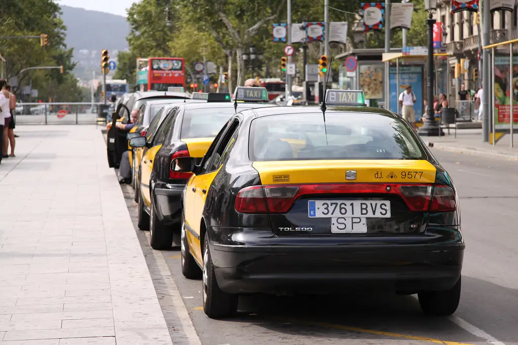 Line of official Barcelona taxis parked along a city avenue, with urban signage and pedestrians nearby.